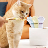 An orange tabby cat licks a small plate held by a human hand, with a white container of colorful cat treats or lick mats nearby. The scene is set in a cozy indoor environment with an orange couch in the background.