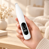 A hand holding a white electric trimmer with a black digital display screen, set against a warm, softly blurred indoor background.